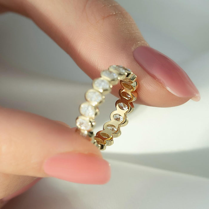 Oval moissanite bezel eternity band held in fingers in yellow gold