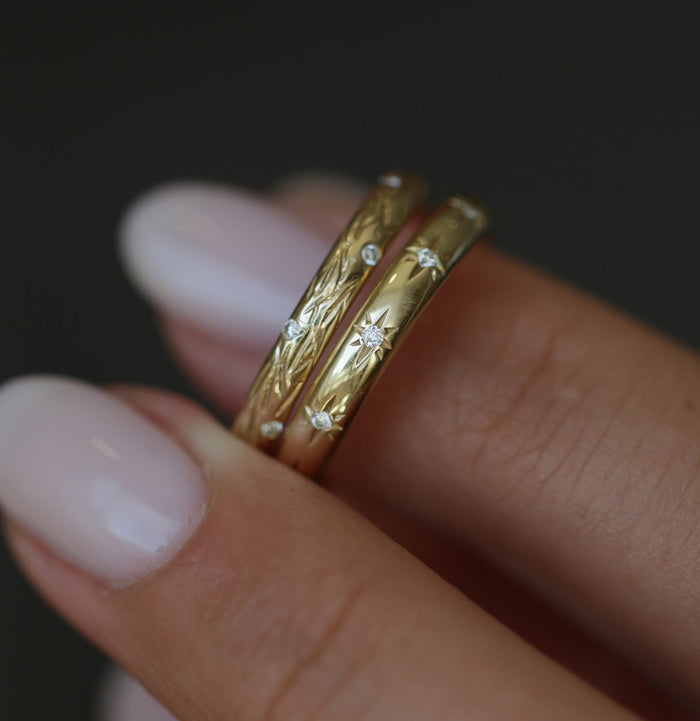 Starburst moissanite wedding band in yellow gold stacked with a leaf textured band, held between fingers