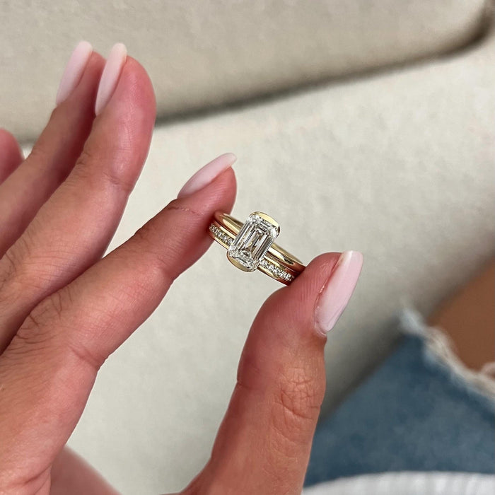 Emerald cut lab grown diamond half bezel solitaire and straight stone set band held between the fingers
