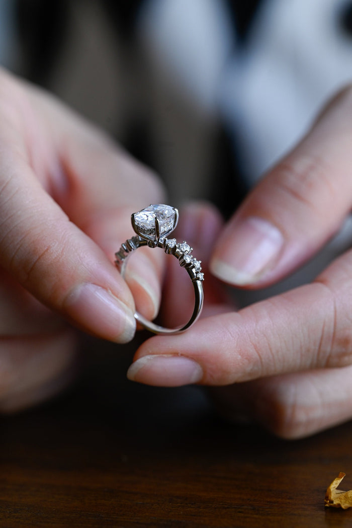Oval moissanite engagement ring with four claws and small round stones set in a staggered zig zag along the shoulders of a slim white gold band, view 2