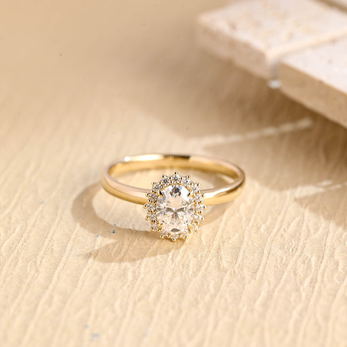 Oval moissanite floral halo engagement ring with a ring of small claw set round stones around the oval on a slim polished yellow gold band