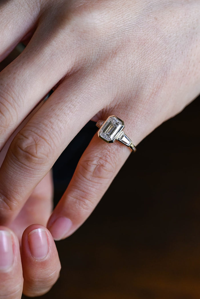 Emerald cut moissanite bezel set three stone engagement ring with a tapered baguette on each side in yellow gold, view 2