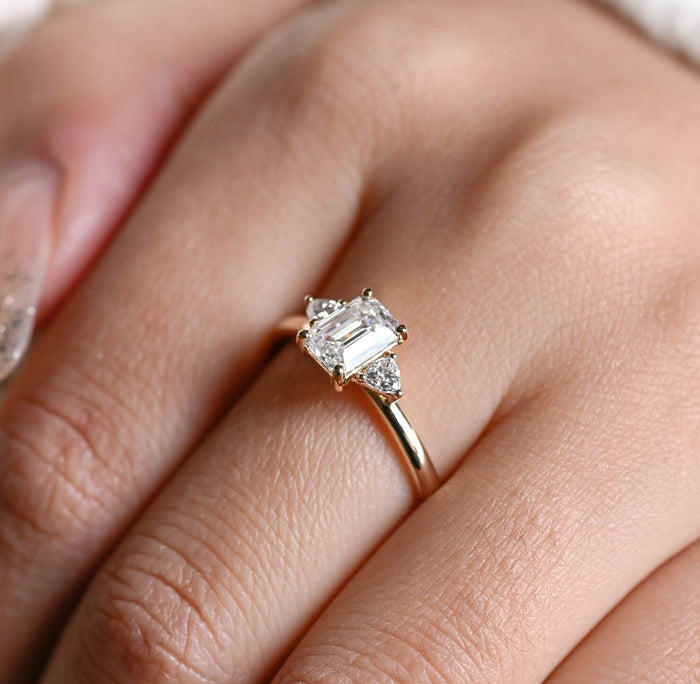Emerald cut moissanite three stone engagement ring with a trillion cut stone on each side of a slim yellow gold band, view 2