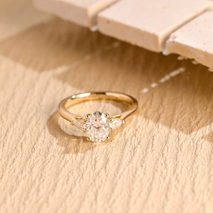 Oval moissanite three stone engagement ring with a pear shaped stone on each side on a plain polished tapered yellow gold band