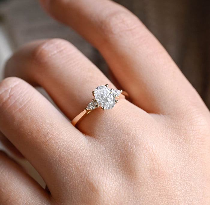 Oval moissanite three stone engagement ring with a pear shaped stone on each side on a plain polished tapered yellow gold band, view 2