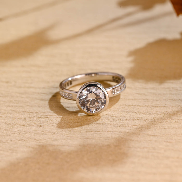 Round moissanite bezel set solitaire engagement ring with a smooth full bezel and a white gold band channel set with small round stones