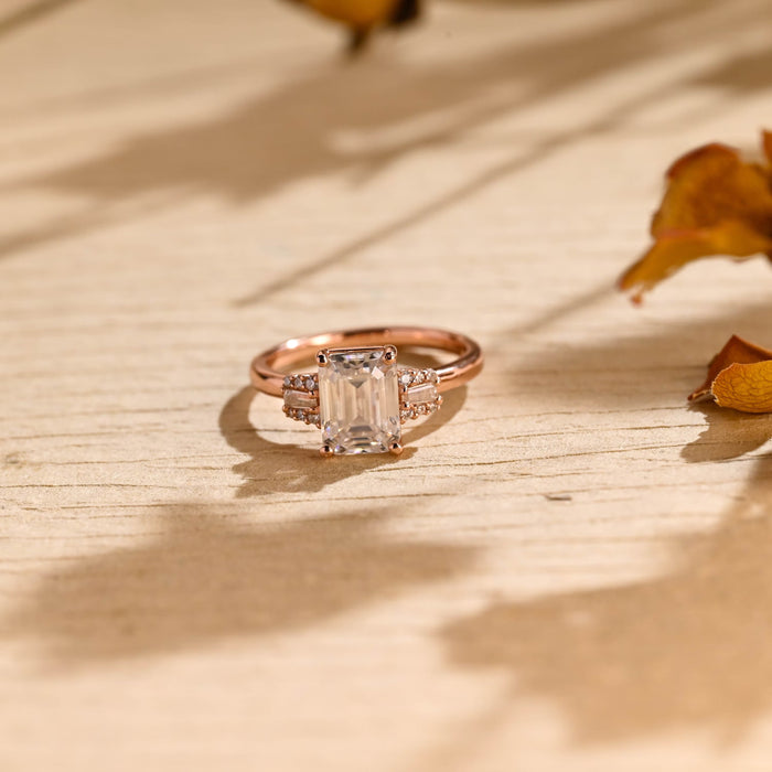 Emerald cut moissanite engagement ring with a baguette framed by tiny round stones on each side of a rose gold band