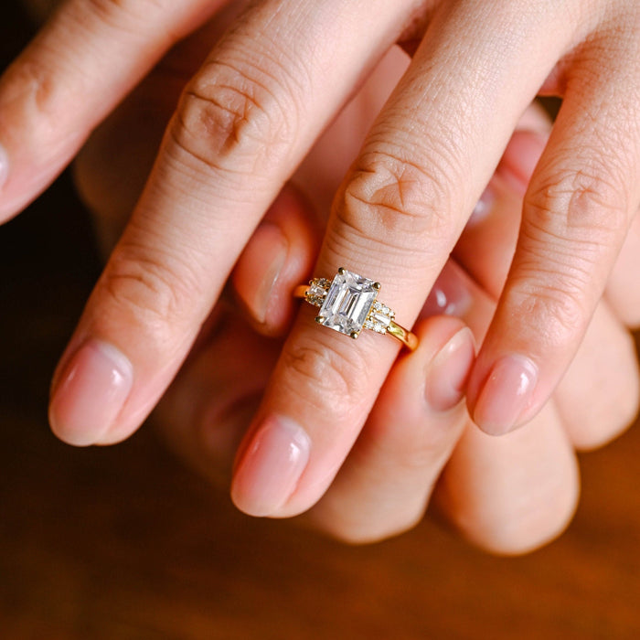 Emerald cut moissanite engagement ring with a baguette framed by tiny round stones on each side of a rose gold band, view 2