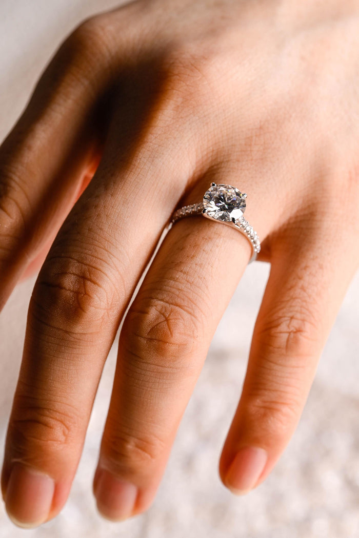 Round moissanite solitaire engagement ring in four slim claws on a fine white gold band pave set with tiny round stones along the shoulders, view 2