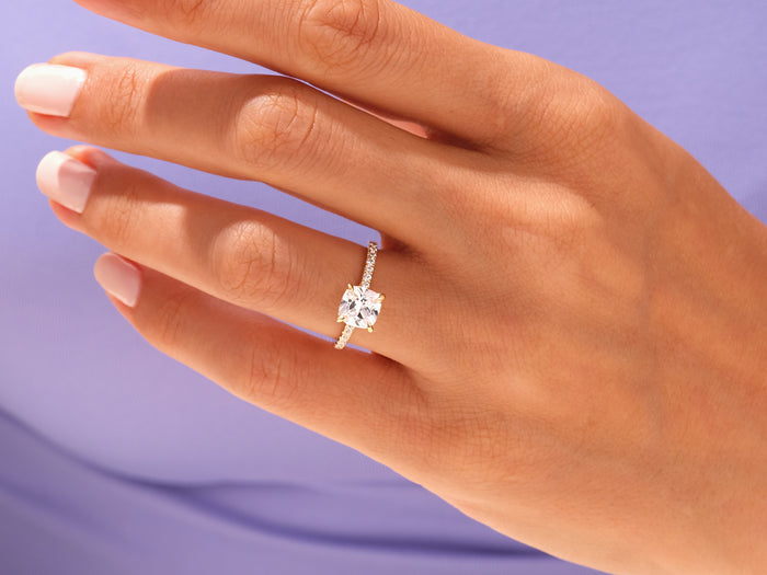 Cushion moissanite scalloped pave engagement ring worn on the hand against a lilac background