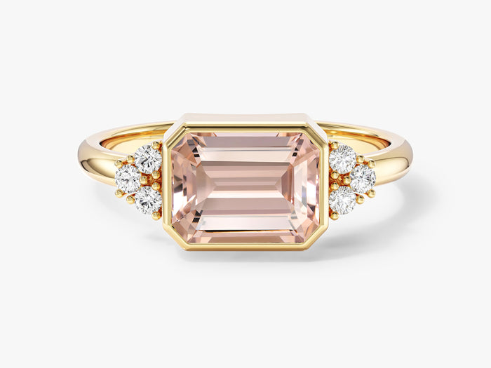 East west emerald cut moissanite bezel engagement ring with round cluster accents in yellow gold, front view, shown with a peach sample center stone