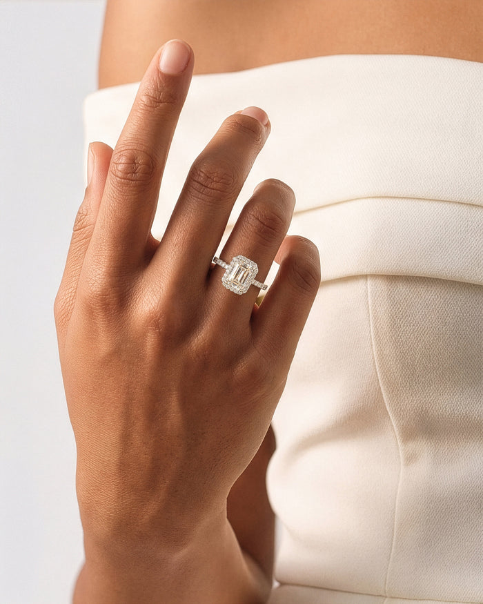 Emerald cut lab grown diamond halo ring in white gold worn on a raised hand against a cream top