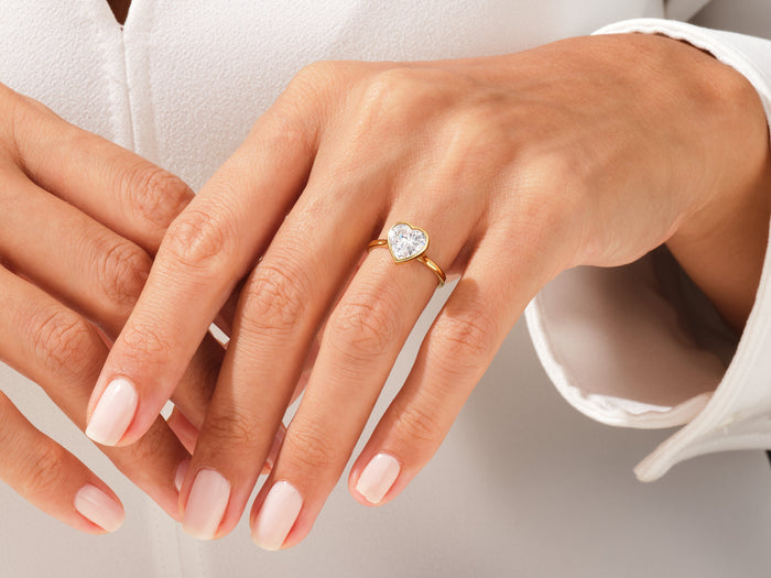 Heart shaped lab grown diamond full bezel ring in yellow gold on crossed hands against a white shirt