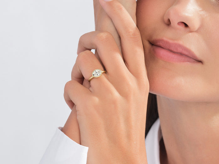 East west oval moissanite bezel ring in yellow gold worn on the hand near the face