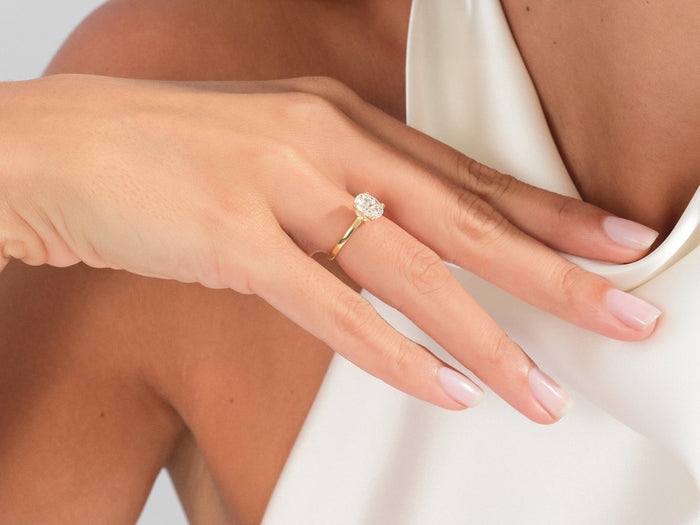 Oval lab grown diamond solitaire in yellow gold worn on a hand resting on an ivory satin neckline