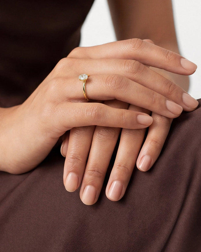 Oval lab grown diamond four prong hidden halo ring in yellow gold on a hand resting on brown fabric