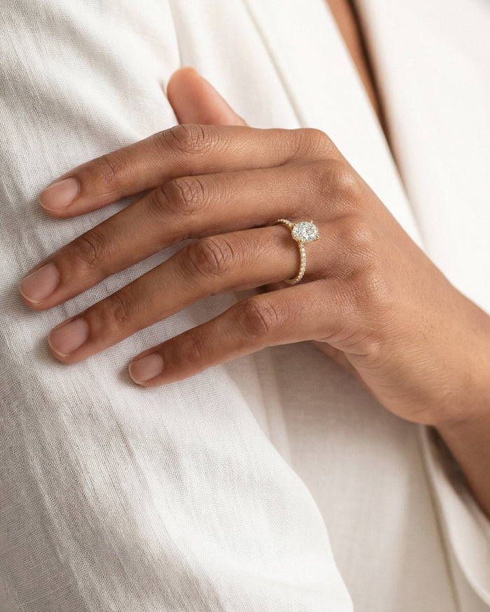 Oval moissanite four claw pave ring in yellow gold worn on the hand against a white jacket