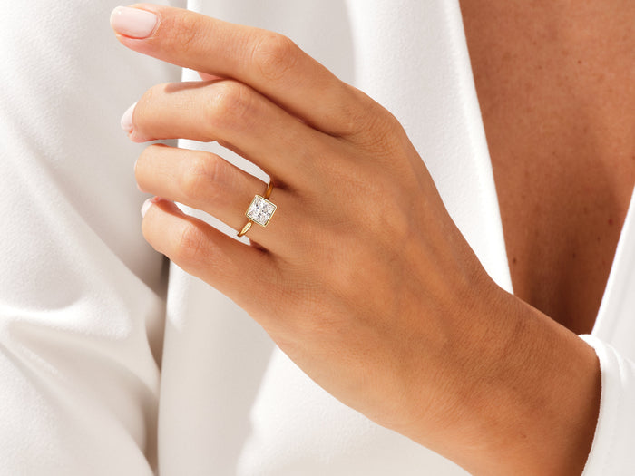 Princess cut lab grown diamond bezel solitaire in yellow gold worn on the hand against a white shirt