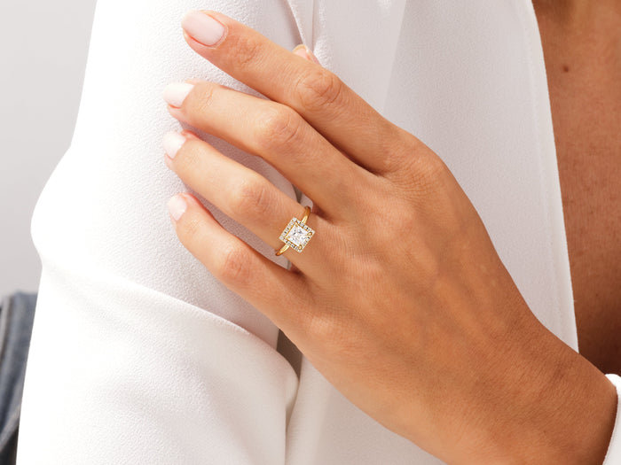 Square princess lab grown diamond halo ring in yellow gold worn on a hand against a white jacket