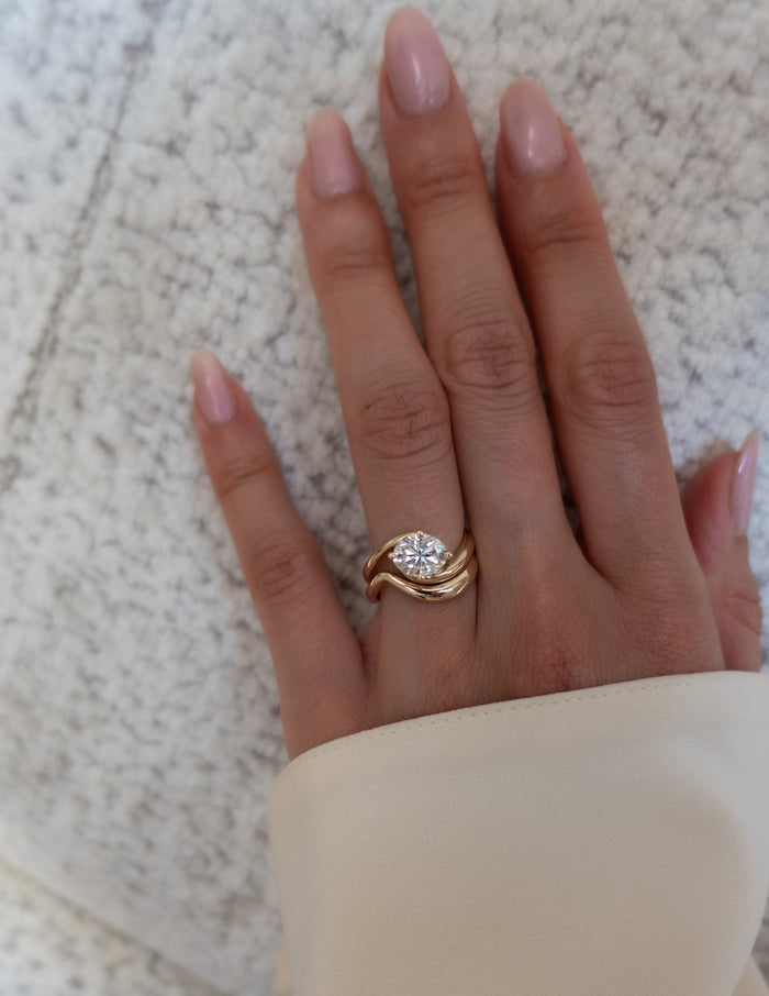 Oval moissanite bypass engagement ring and plain wave wedding band on a hand resting on a textured throw