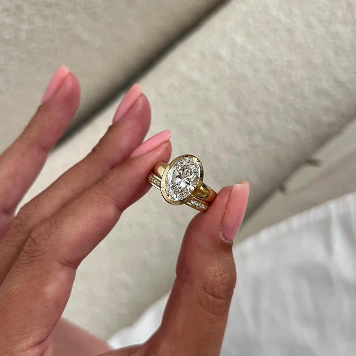 Oval moissanite full bezel engagement ring and pave band held between the fingers