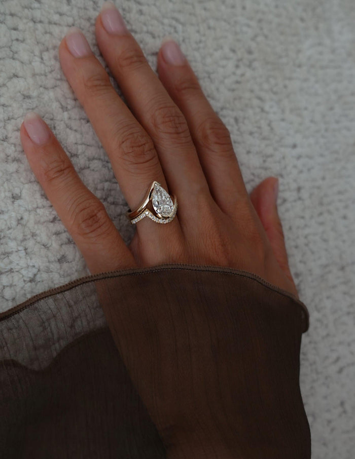 Pear lab grown diamond bypass engagement ring and curved pave wedding band on a hand resting on a textured throw