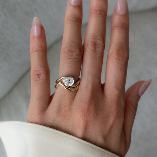 East-west pear moissanite swirl bridal set with plain wave band in yellow gold worn on the hand