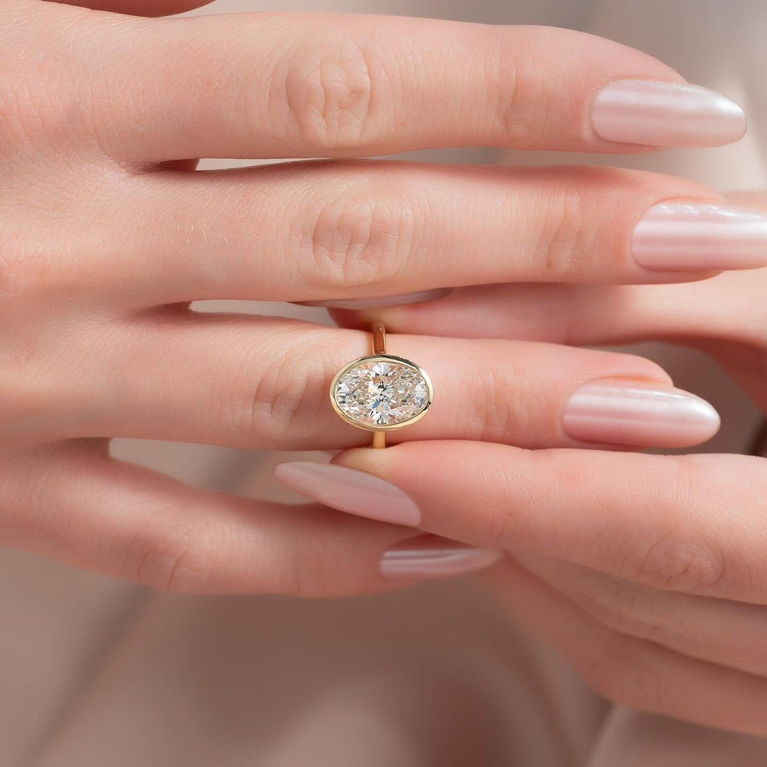 East west oval lab grown diamond bezel ring on a slim yellow gold band worn on the hand