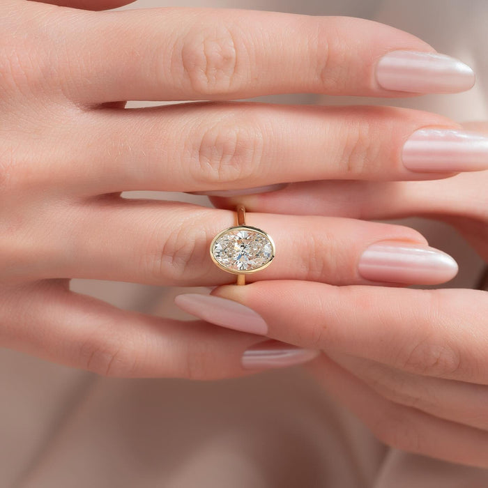 East west oval lab grown diamond bezel ring on a slim yellow gold band worn on the hand