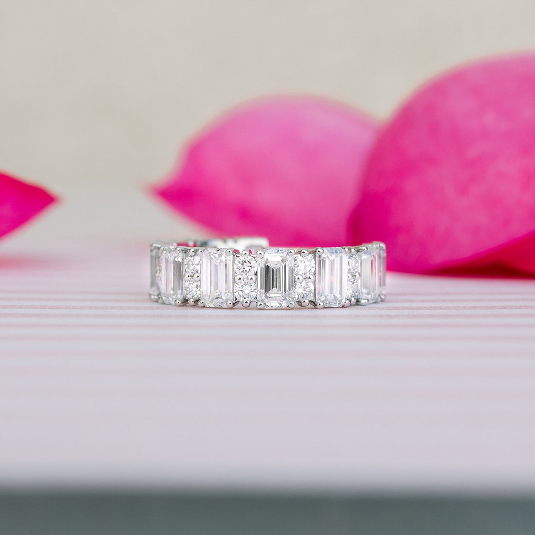 White gold emerald cut and round moissanite three quarter eternity band standing on a striped surface in front of pink petals
