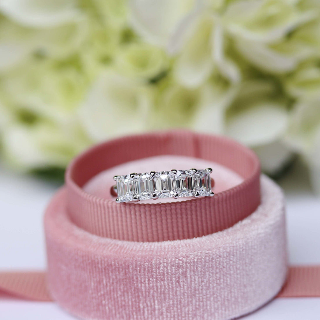 White gold five stone emerald cut moissanite band in a pink velvet ring box in front of white flowers
