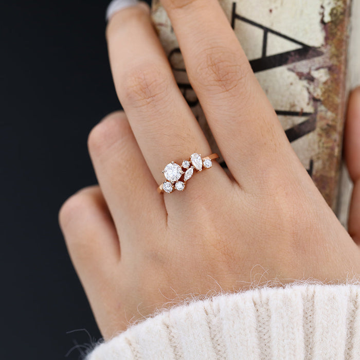 Rose gold cluster moissanite ring with a larger round stone and marquise and pear stones worn on a finger holding a book