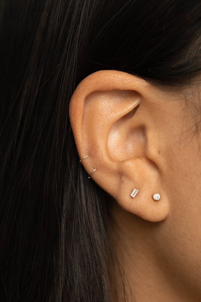 Ear wearing a baguette lab grown diamond bezel stud next to a small round stud, styled with thin gold hoops