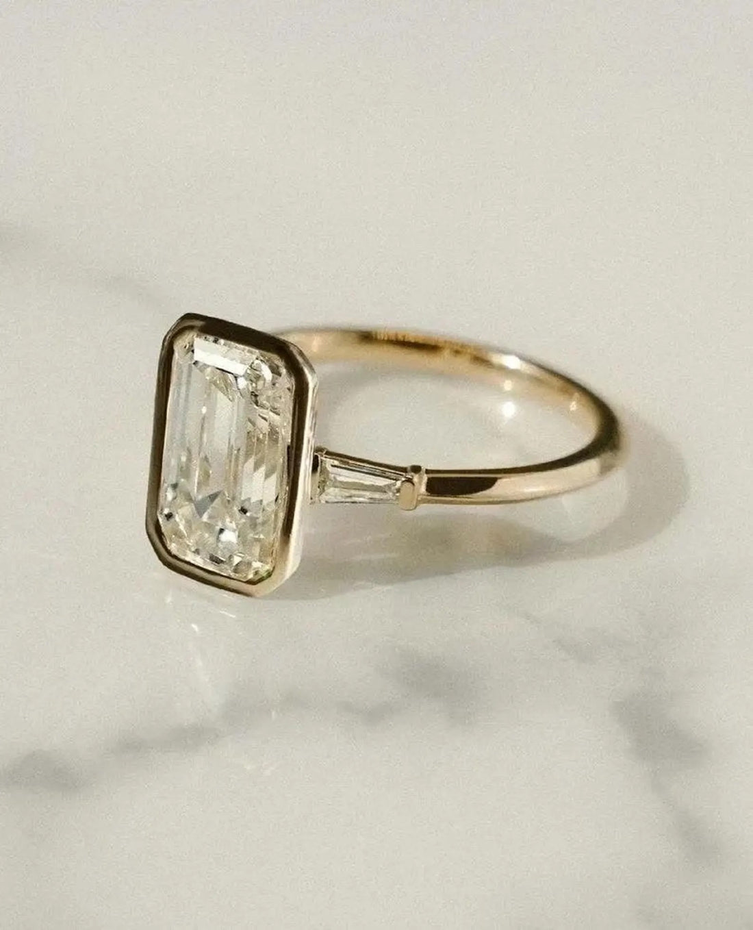 Bezel set emerald cut lab grown diamond ring with tapered baguette shoulders in yellow gold