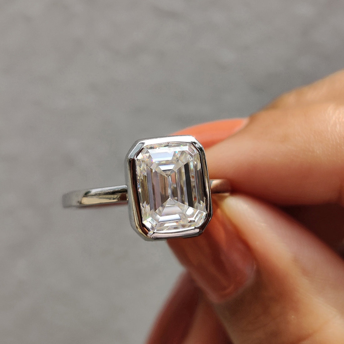 Emerald cut lab grown diamond bezel solitaire engagement ring in white gold held between two fingers