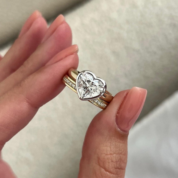 Heart lab grown diamond bezel solitaire and pave wedding band held between the fingers