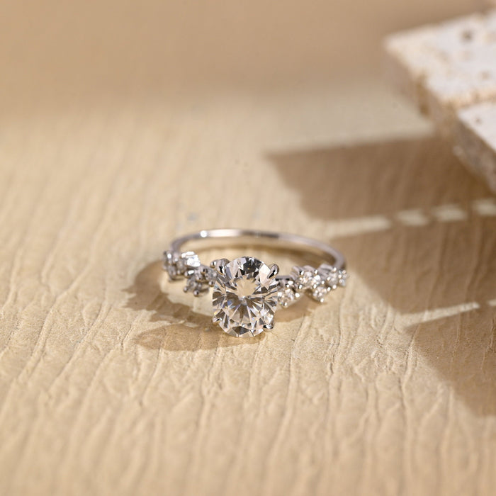 Oval moissanite engagement ring with four claws and small round stones set in a staggered zig zag along the shoulders of a slim white gold band