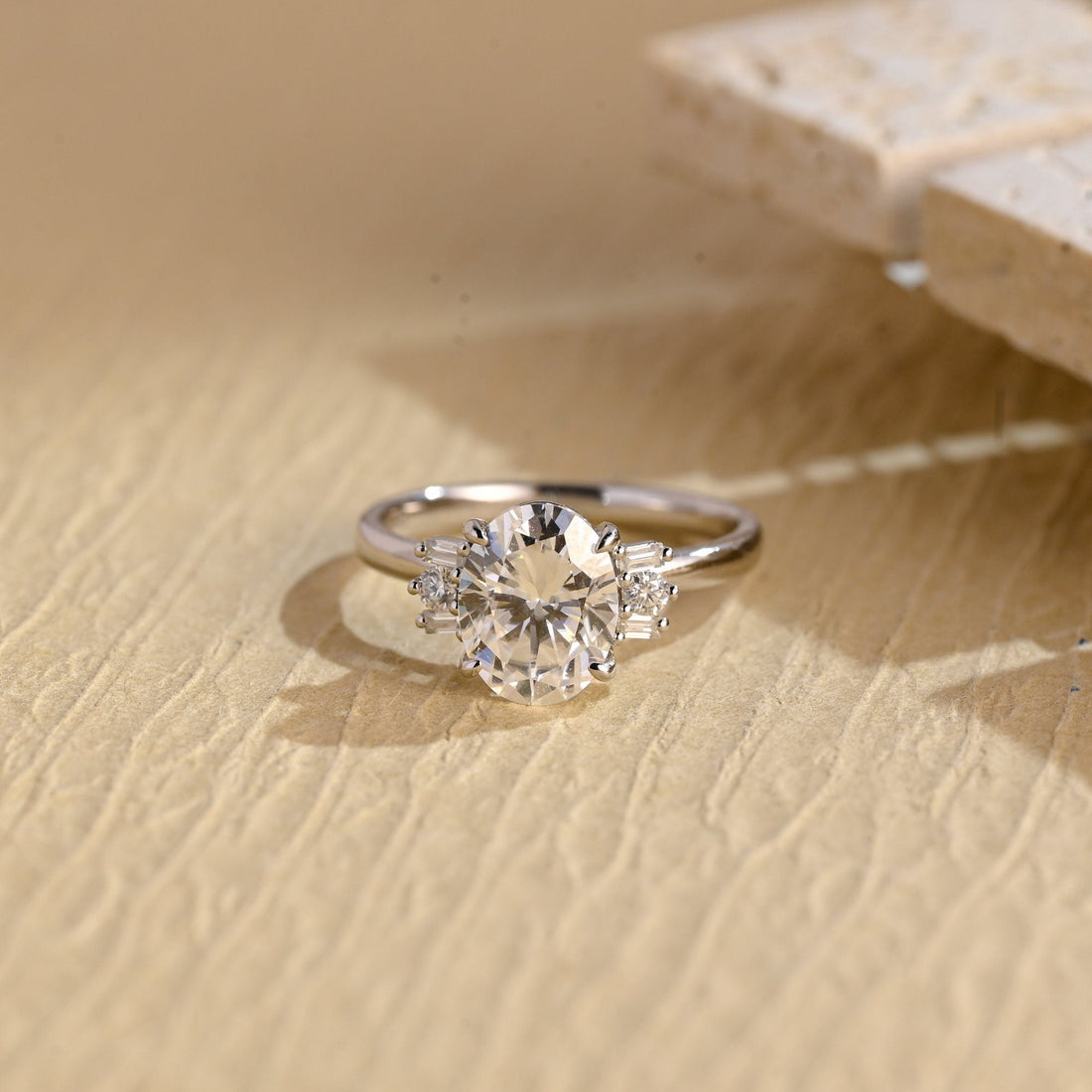 Oval moissanite engagement ring with two baguette stones and a round stone stacked on each side of the oval on a plain polished white gold band