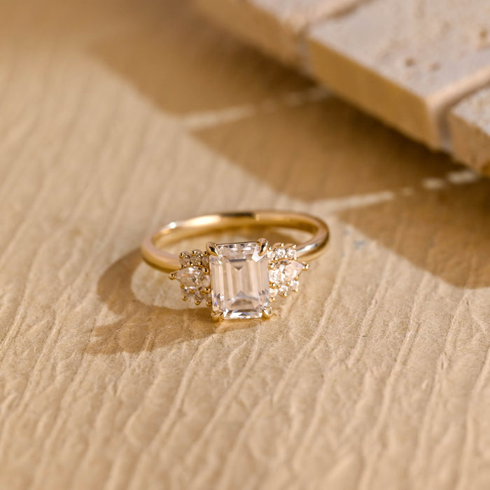 Emerald cut moissanite engagement ring with a pear and round stone cluster on each side of a plain yellow gold band