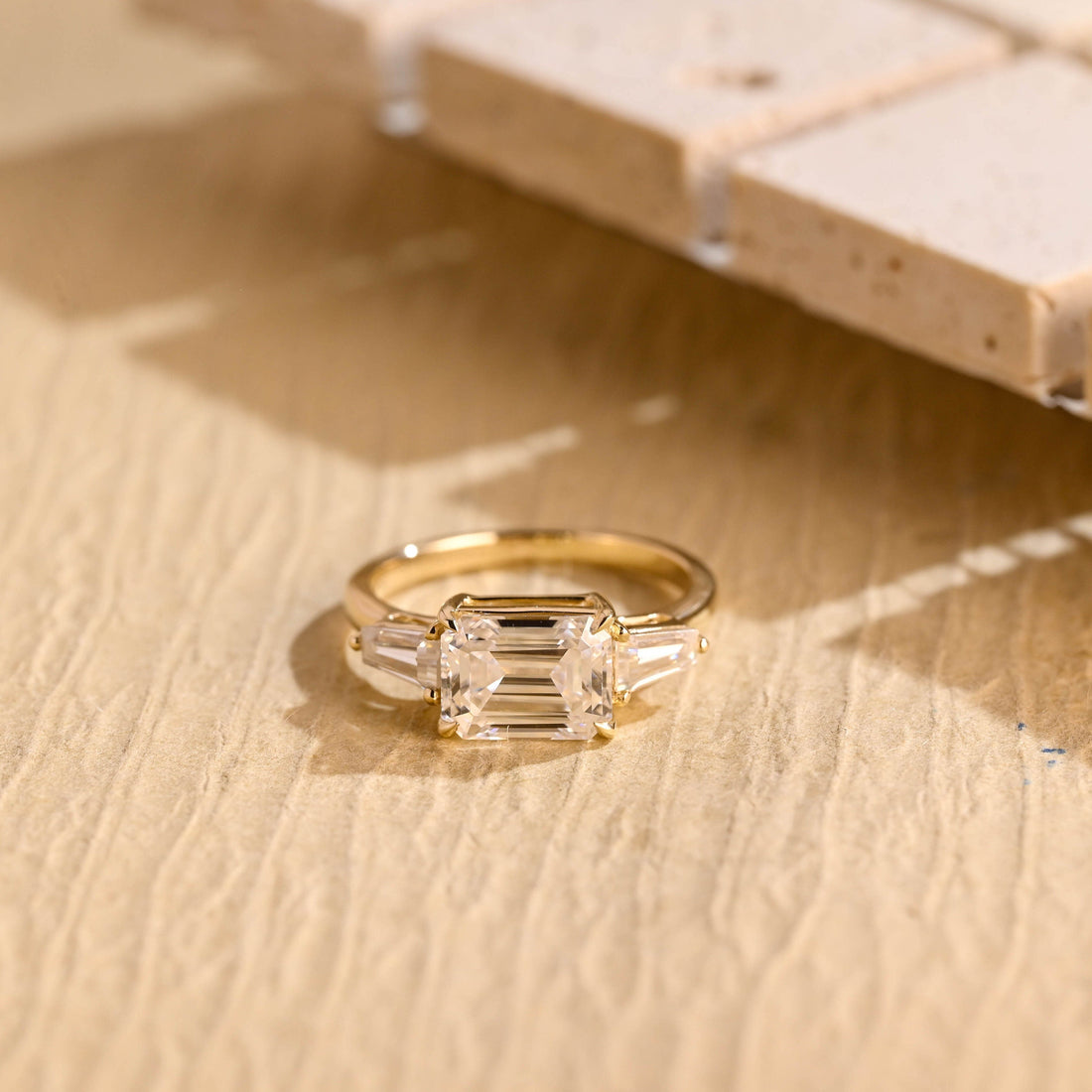 East west emerald cut moissanite three stone engagement ring with tapered baguette side stones on a polished yellow gold band