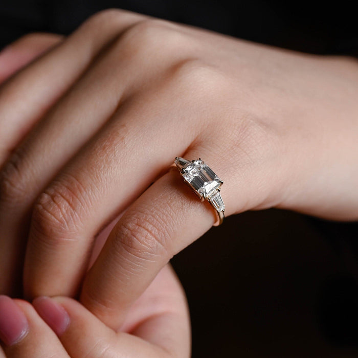 East west emerald cut moissanite three stone engagement ring with tapered baguette side stones on a polished yellow gold band, view 2