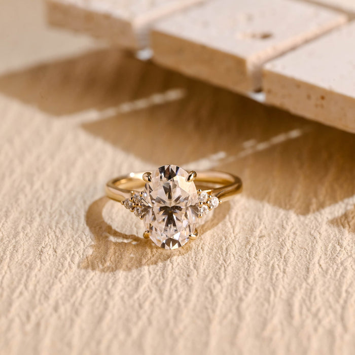 Oval moissanite engagement ring with four claws and a cluster of three small round stones on each side of a plain yellow gold band