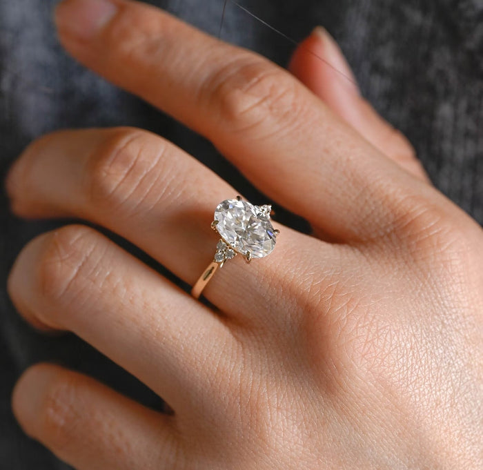 Oval moissanite engagement ring with four claws and a cluster of three small round stones on each side of a plain yellow gold band, view 2