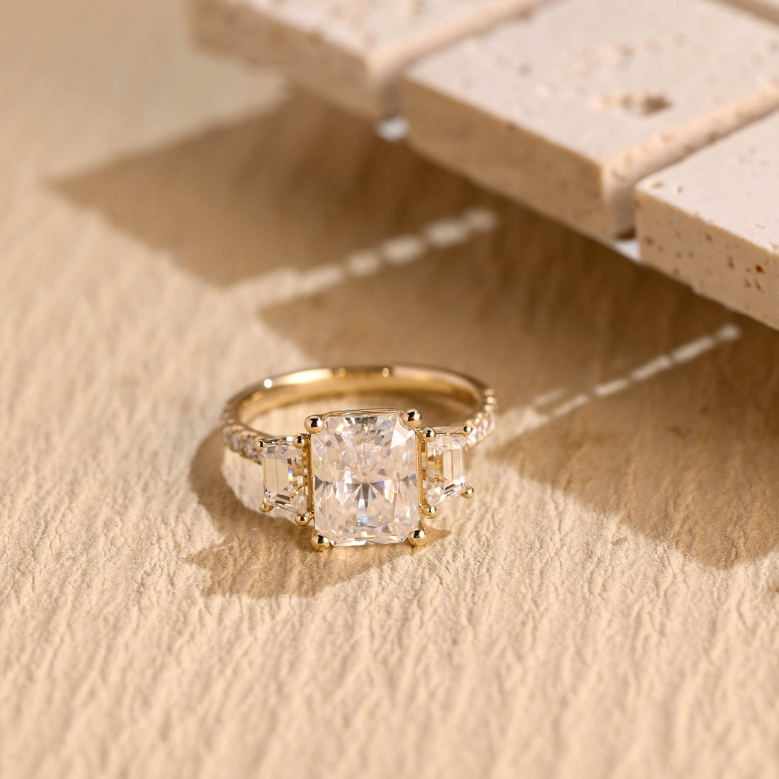 Radiant cut moissanite three stone engagement ring with tapered baguette side stones in claws and a pave set yellow gold band