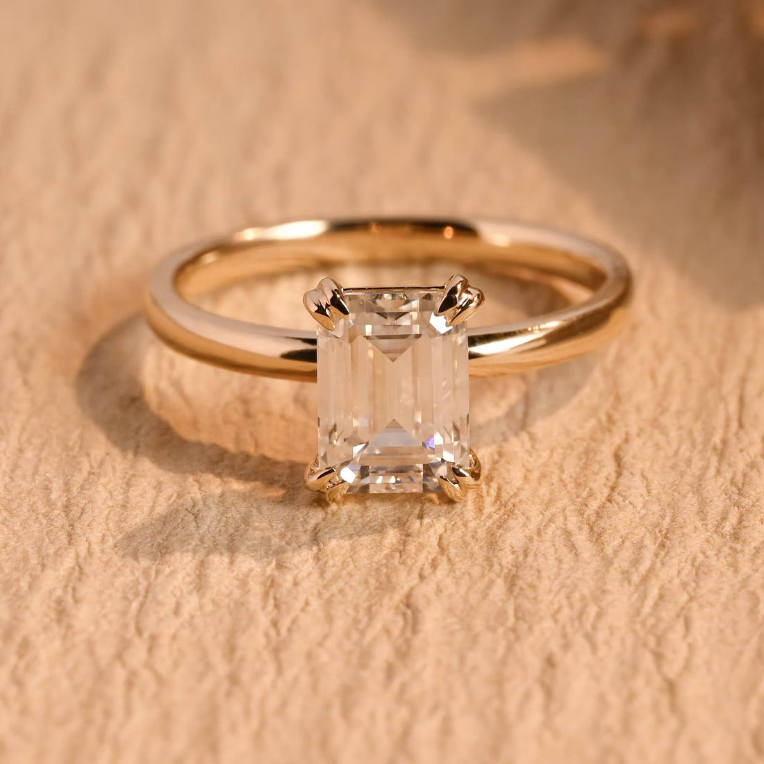 Emerald cut moissanite solitaire engagement ring held by double claws at each corner on a slim polished yellow gold band
