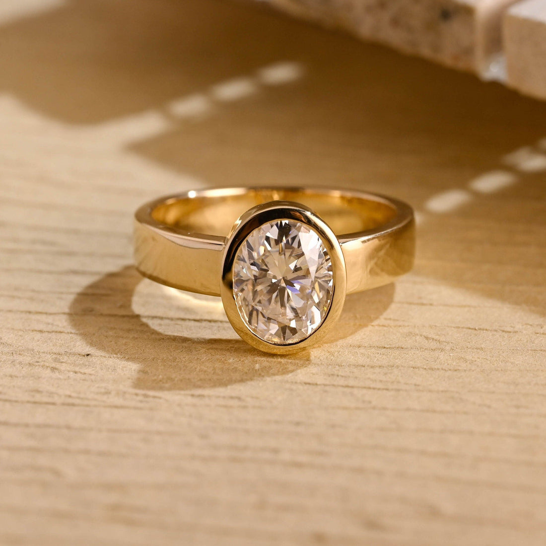 Oval moissanite bezel set solitaire engagement ring on a wide flat polished yellow gold band
