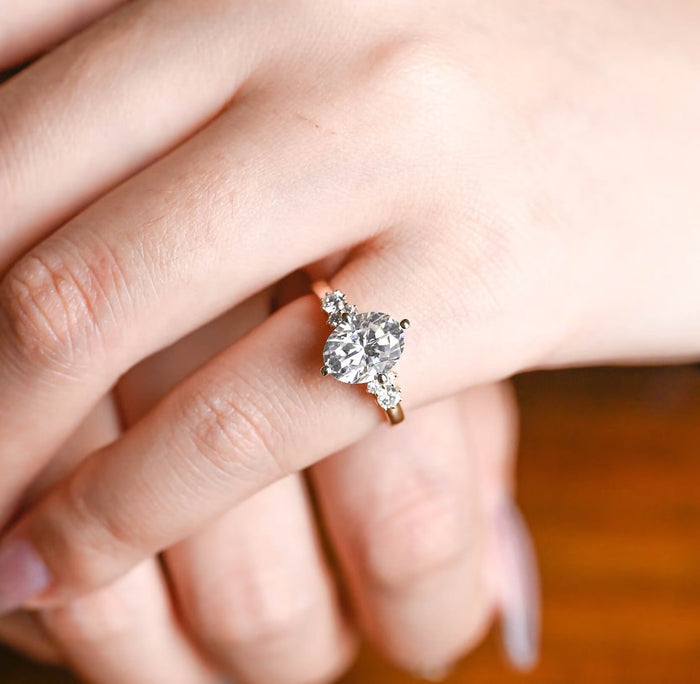 Oval moissanite engagement ring held by four compass point claws with a cluster of small round stones on each side on a slim plain yellow gold band, view 2