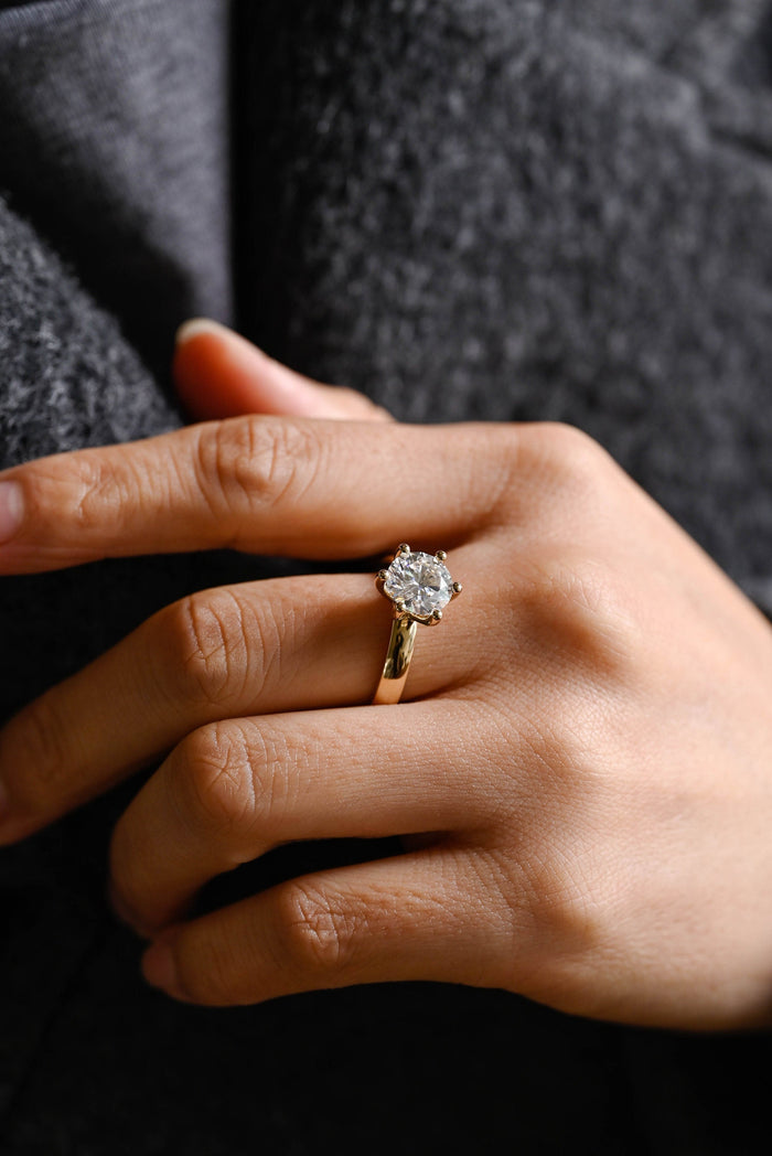 Round moissanite solitaire engagement ring held by six ball tipped claws on a broad polished tapered yellow gold band, view 2