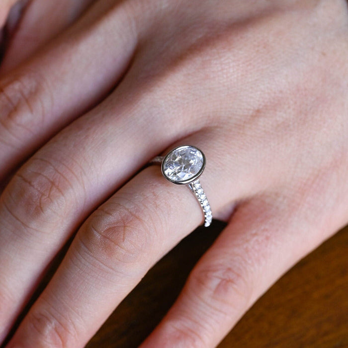 Bezel set oval moissanite engagement ring on a slim white gold band set with small round stones, view 2