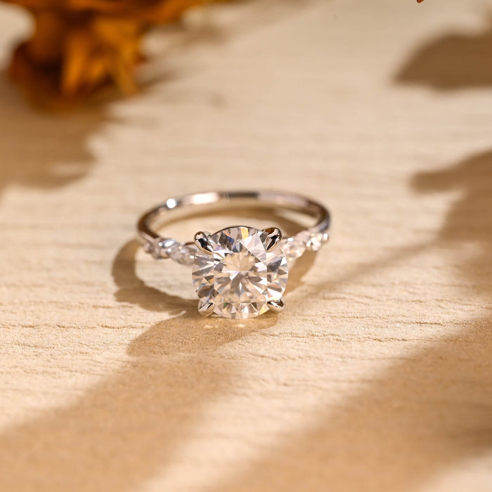 Round moissanite solitaire engagement ring in four claws with a marquise and a small round stone at each shoulder of a slim white gold band